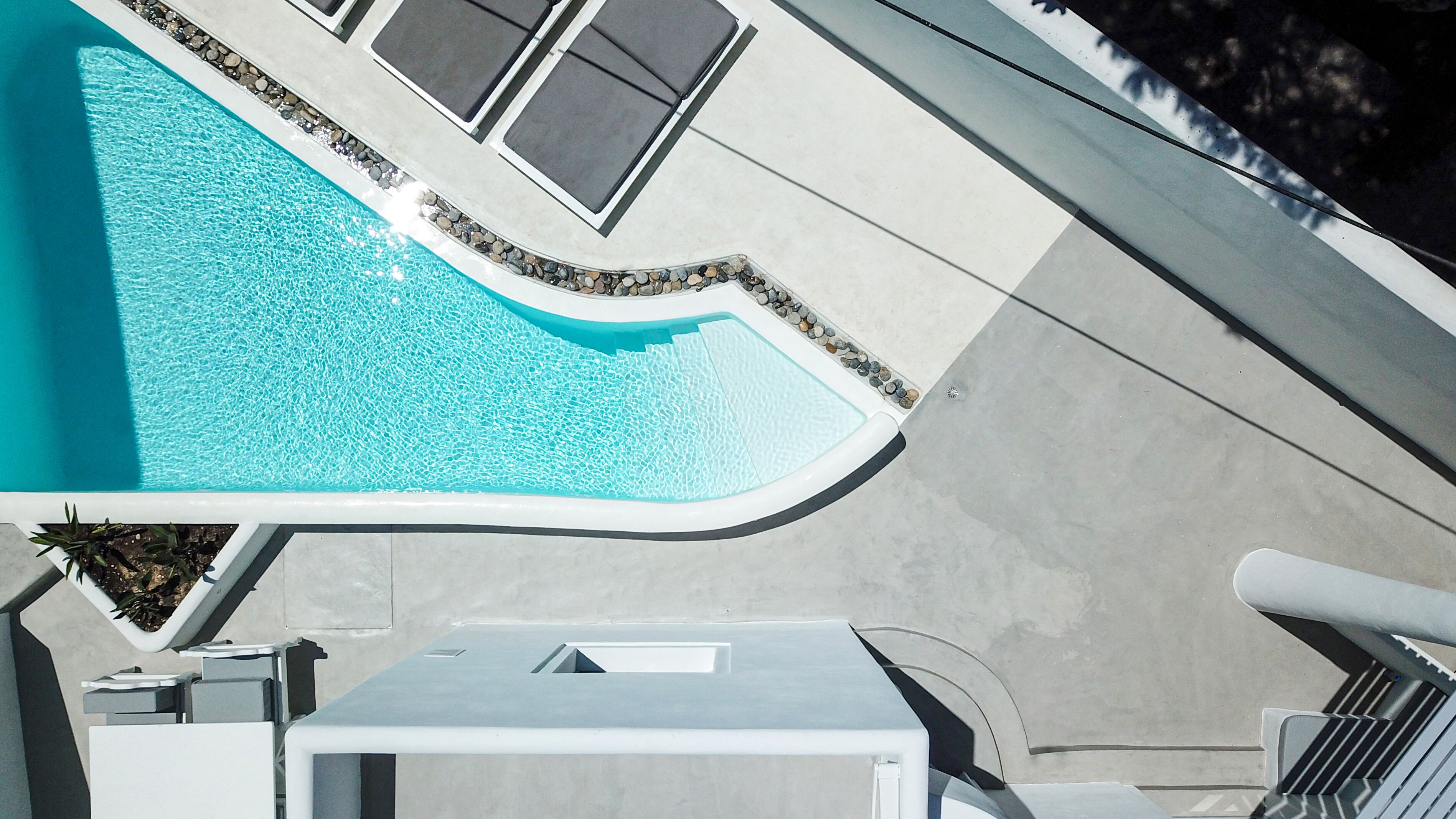 An aerial view of a swimming pool with a curved edge, surrounded by a pebble-lined walkway and adjacent lounge chairs.