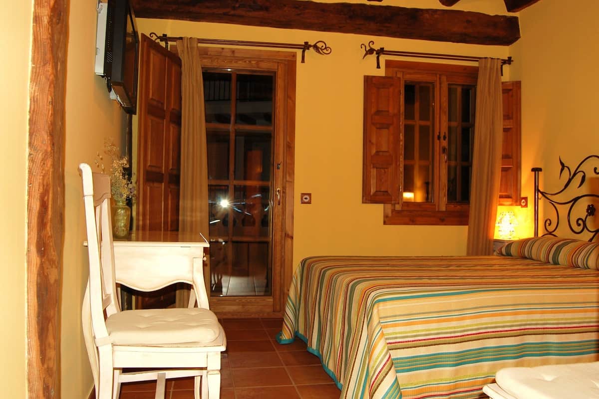 A bedroom with a bed, a chair, a window with curtains, and a door leading to another room.