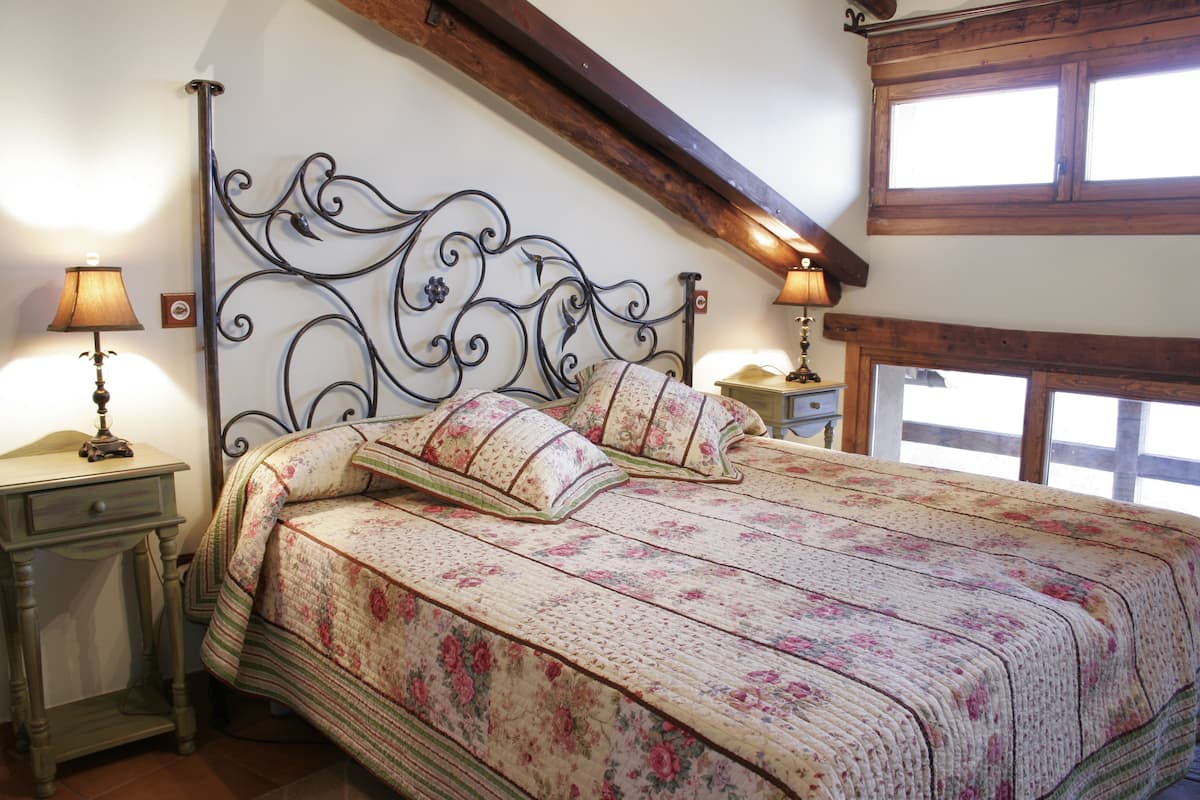 A bedroom with a floral bedspread, a wrought iron headboard, two bedside tables with lamps, and a window with wooden beams.