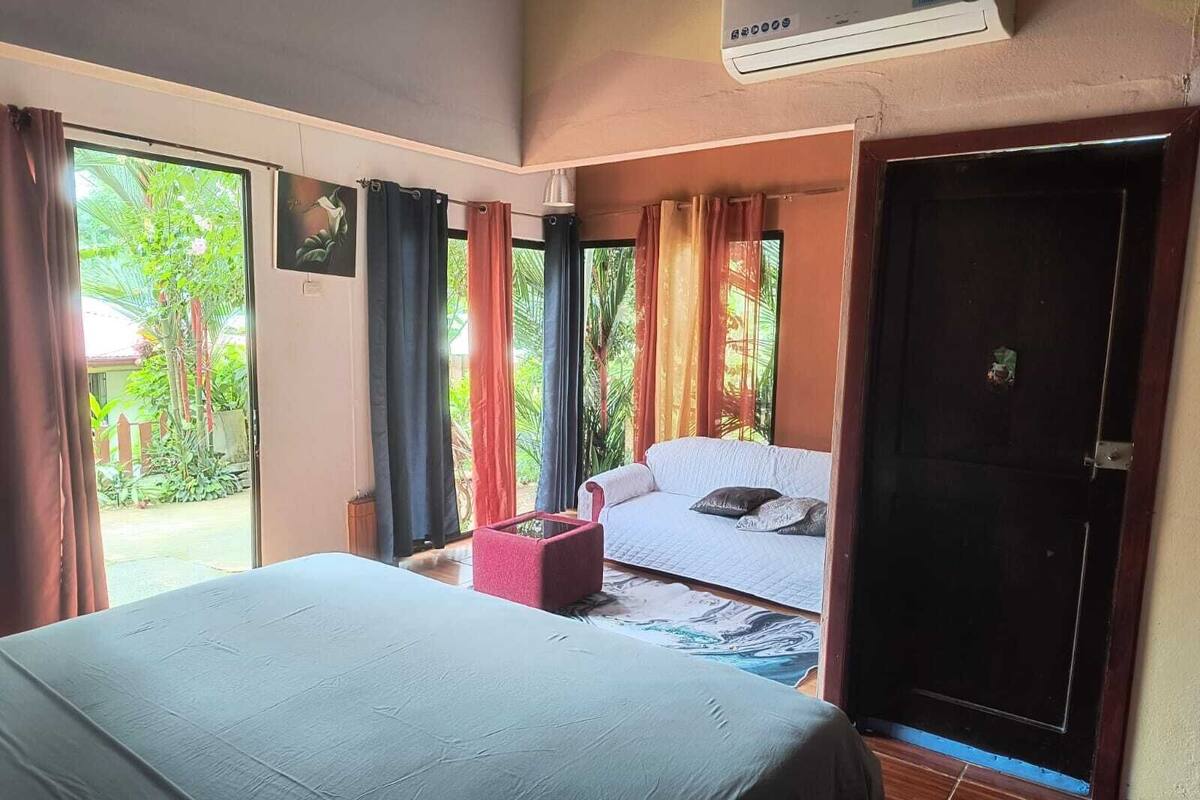 A room with a bed, a sofa, and a small table. There is a door and a window with curtains.
