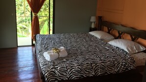 A bedroom with a zebra-patterned bedspread, a wooden headboard, a bedside lamp, and a view of greenery through a window with curtains.