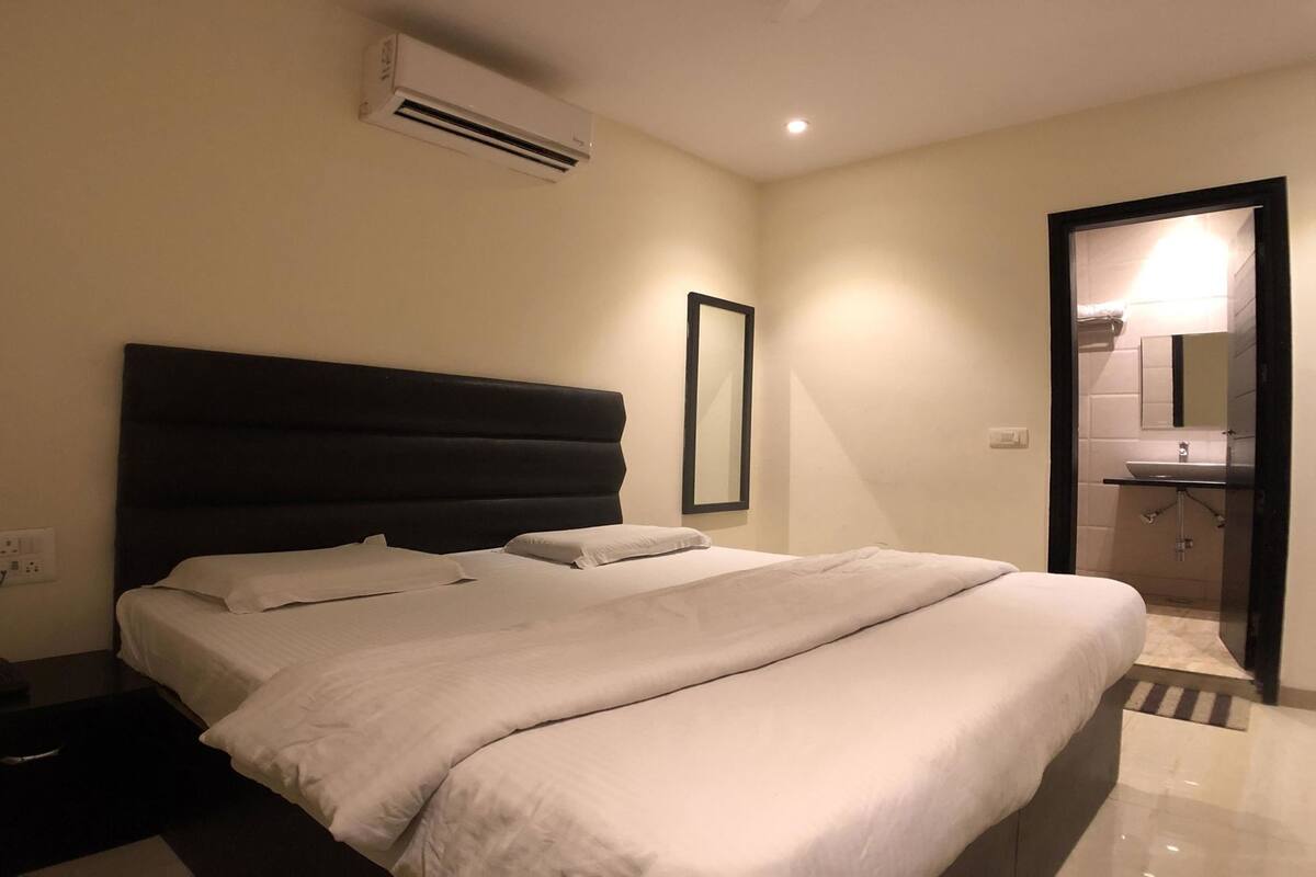 A hotel room with a bed, a nightstand, an air conditioning unit, a mirror, and a bathroom visible through an open door.