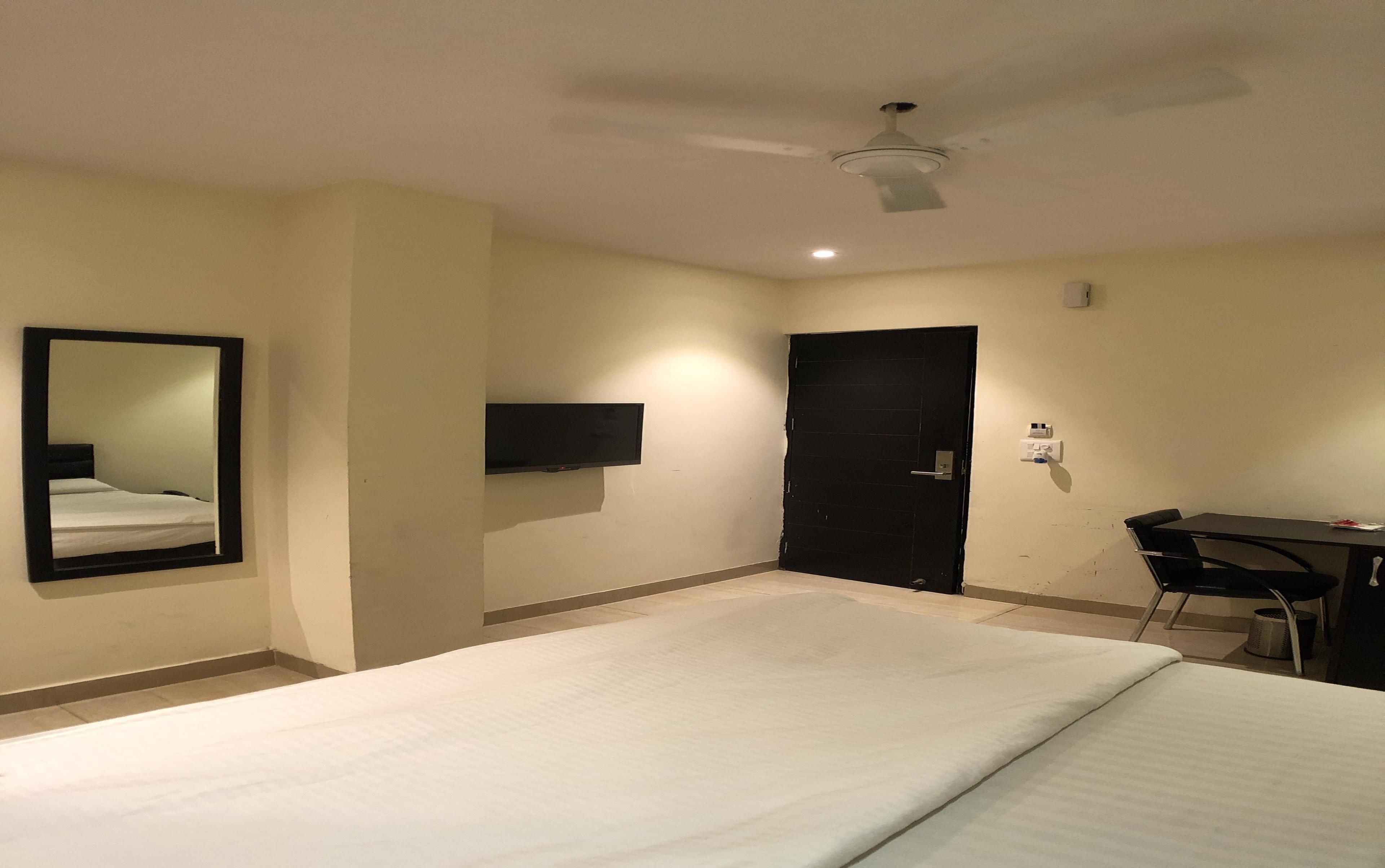A hotel room with a bed, a desk, a chair, a door, a ceiling fan, and a TV mounted on the wall.