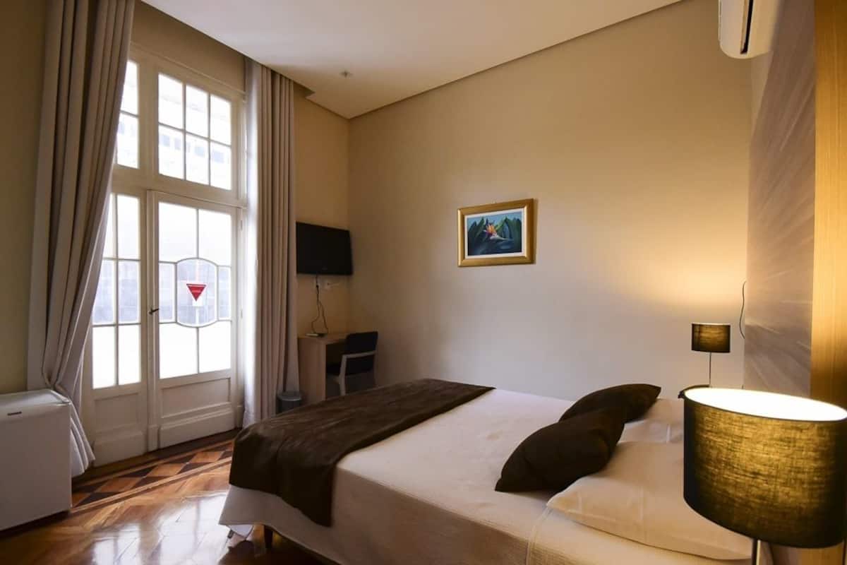 Une chambre d’hôtel avec un lit, un bureau, une chaise, une télévision et une fenêtre avec des rideaux.
