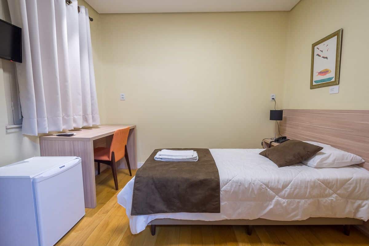Une chambre d’hôtel équipée d’un lit, d’un bureau, d’une chaise, d’un petit réfrigérateur et d’un téléviseur fixé au mur.