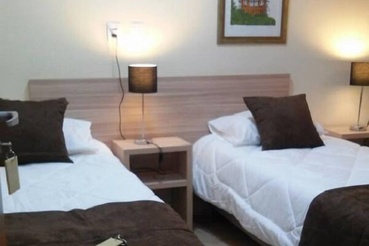 Une chambre d’hôtel avec deux lits, chacun doté d’une tête de lit marron, de draps blancs et d’une petite table de chevet avec une lampe. Un tableau encadré est accroché au mur au-dessus des lits.