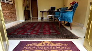 A hotel entrance with a red patterned rug, a purple doormat with 'Hotel Praca da Malizia' text, a blue chair, and a wooden desk with a computer. - Hotel Praça da Matriz (Porto Alegre)