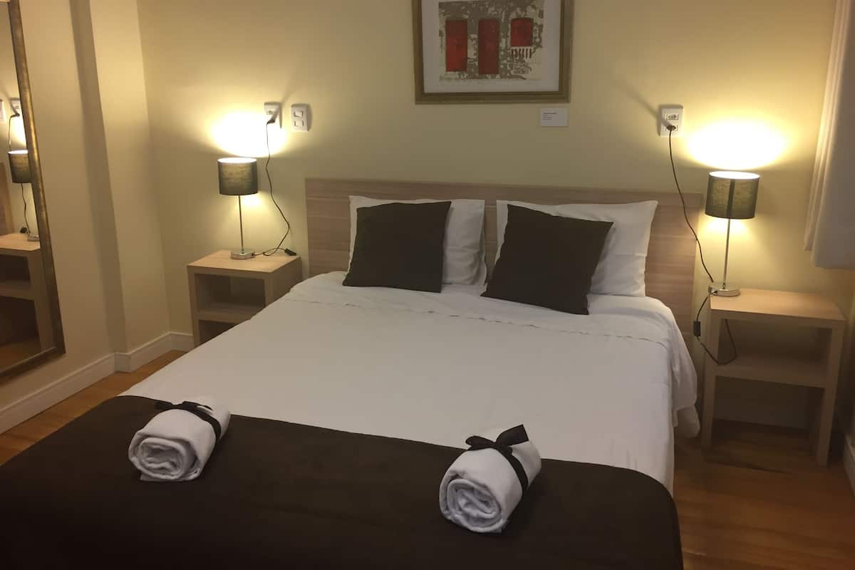 Une chambre d’hôtel avec un grand lit, deux tables de chevet avec des lampes, un tableau encadré au mur et une serviette roulée posée sur le lit.