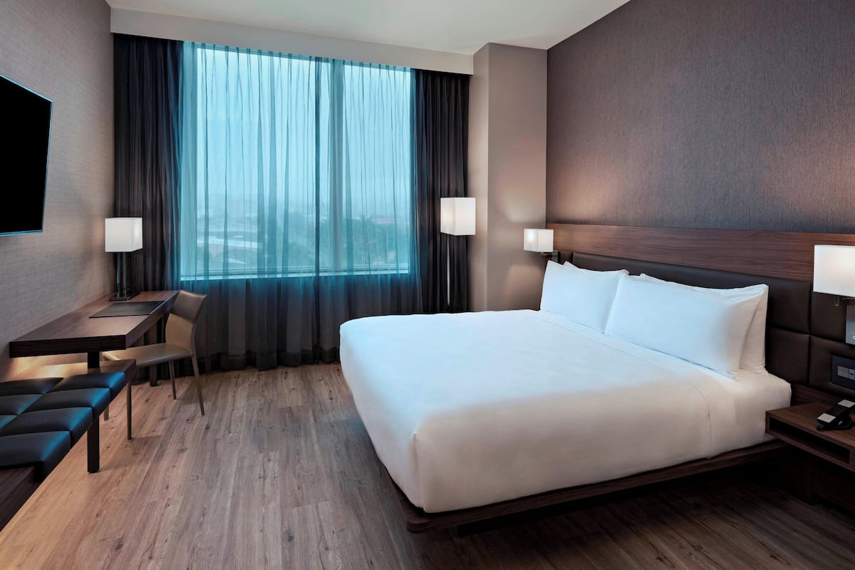 Quarto de hotel com uma cama grande, uma mesa com cadeira, uma televisão na parede e uma janela com cortinas.