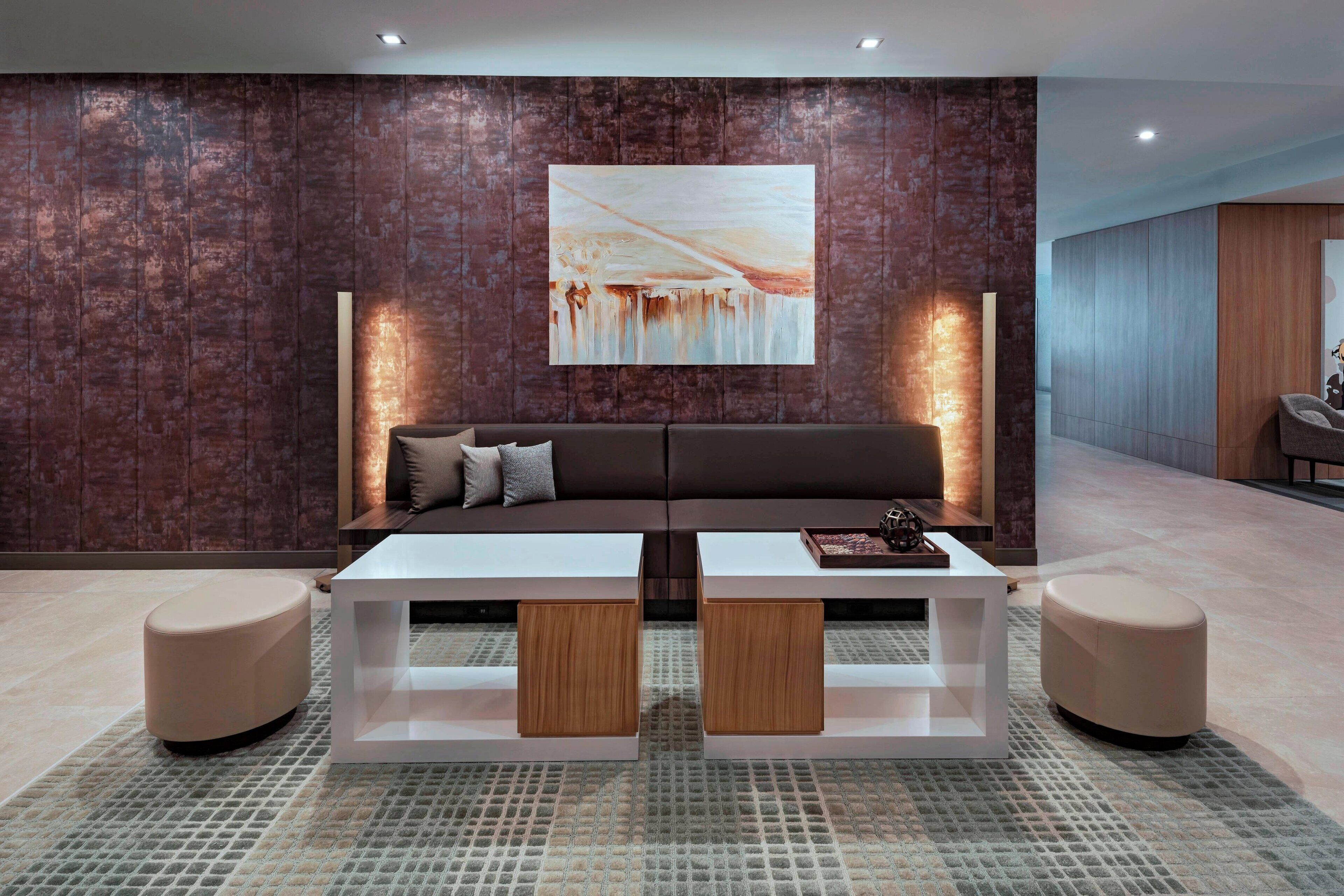A modern living room with a brown textured wall, a large abstract painting, a brown sofa, a white coffee table, and two ottomans.