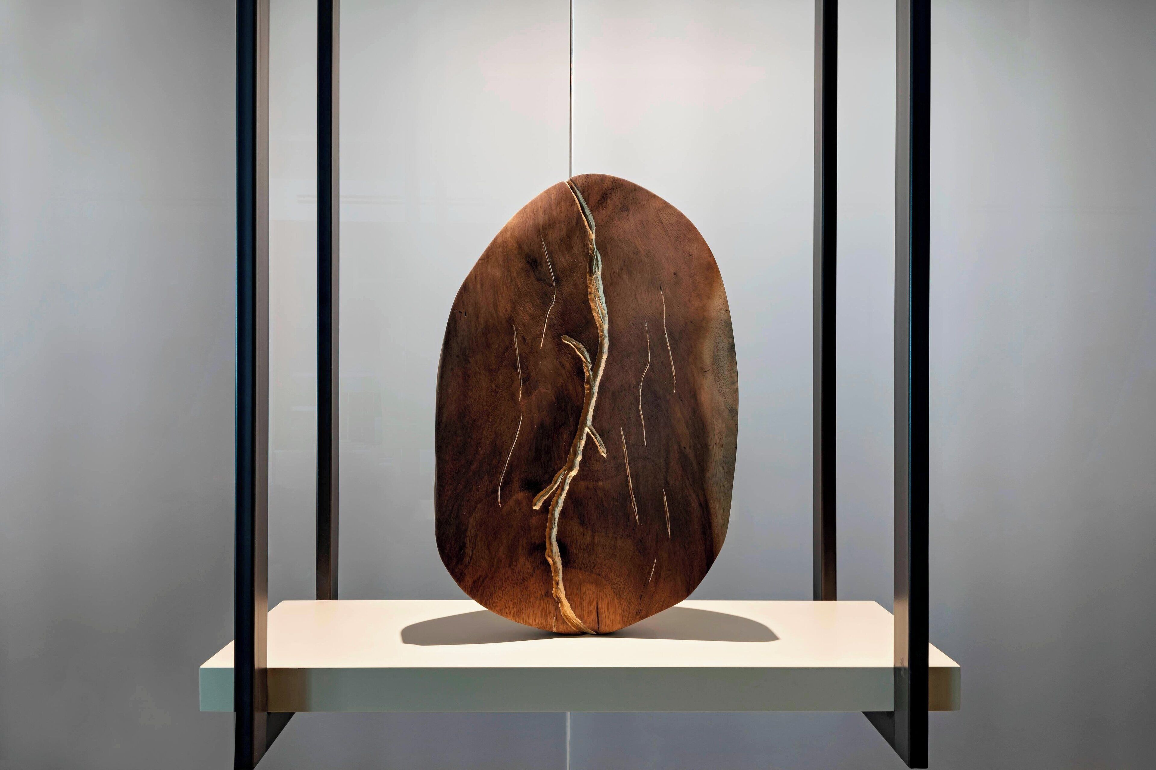 Una gran escultura de madera ovalada con una grieta visible, expuesta sobre un pedestal blanco frente a un fondo neutro.