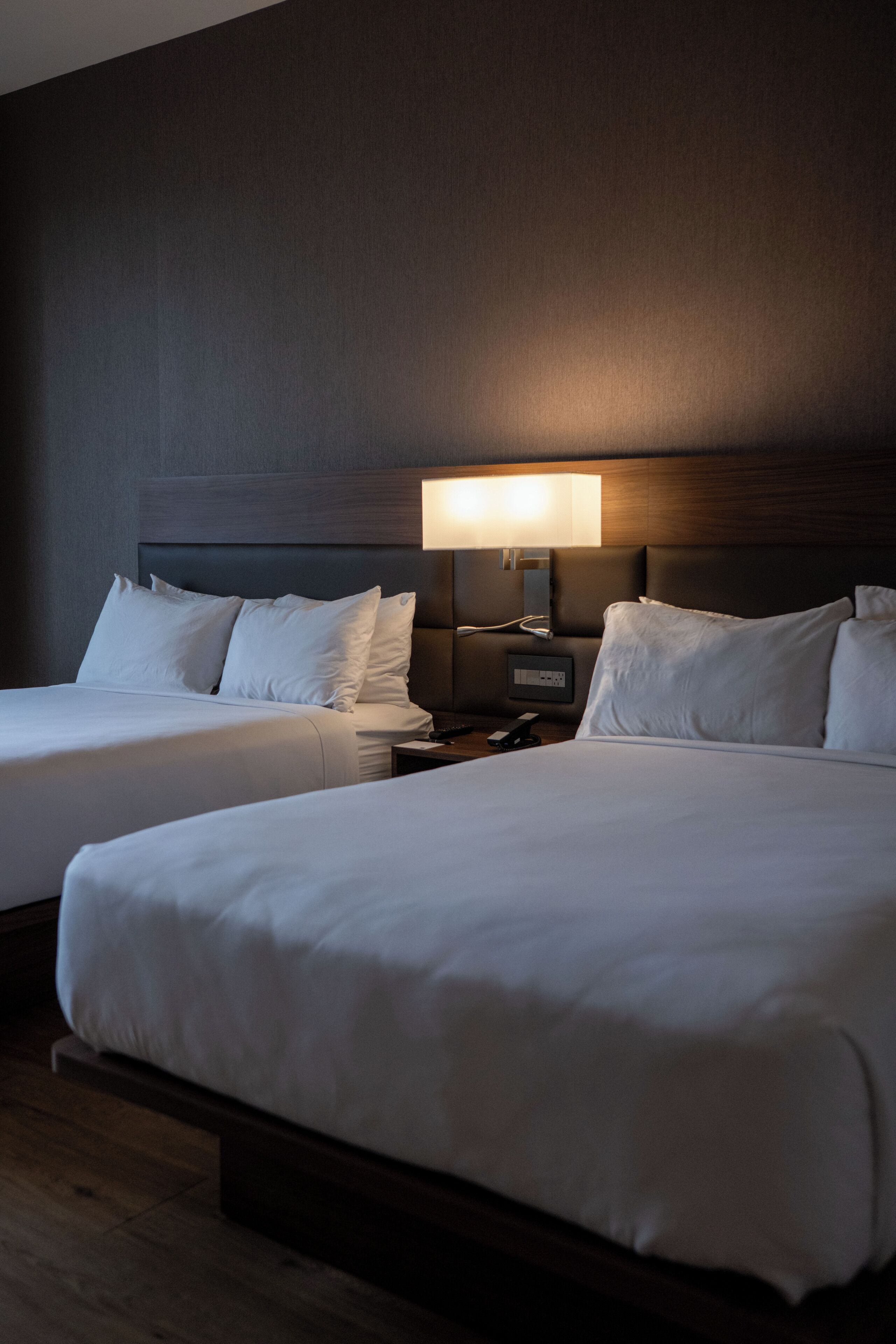 A hotel room with two beds, a nightstand, and a wall-mounted lamp.