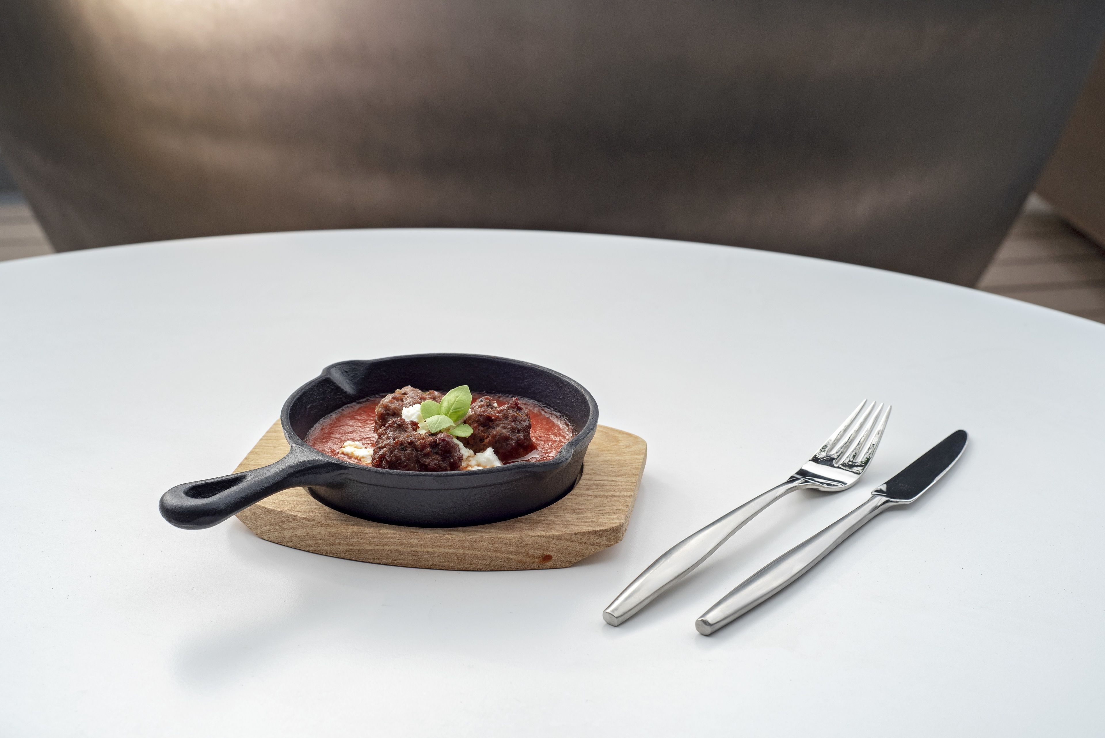 A black skillet with meatballs on a wooden board, accompanied by a fork and knife on a white table.