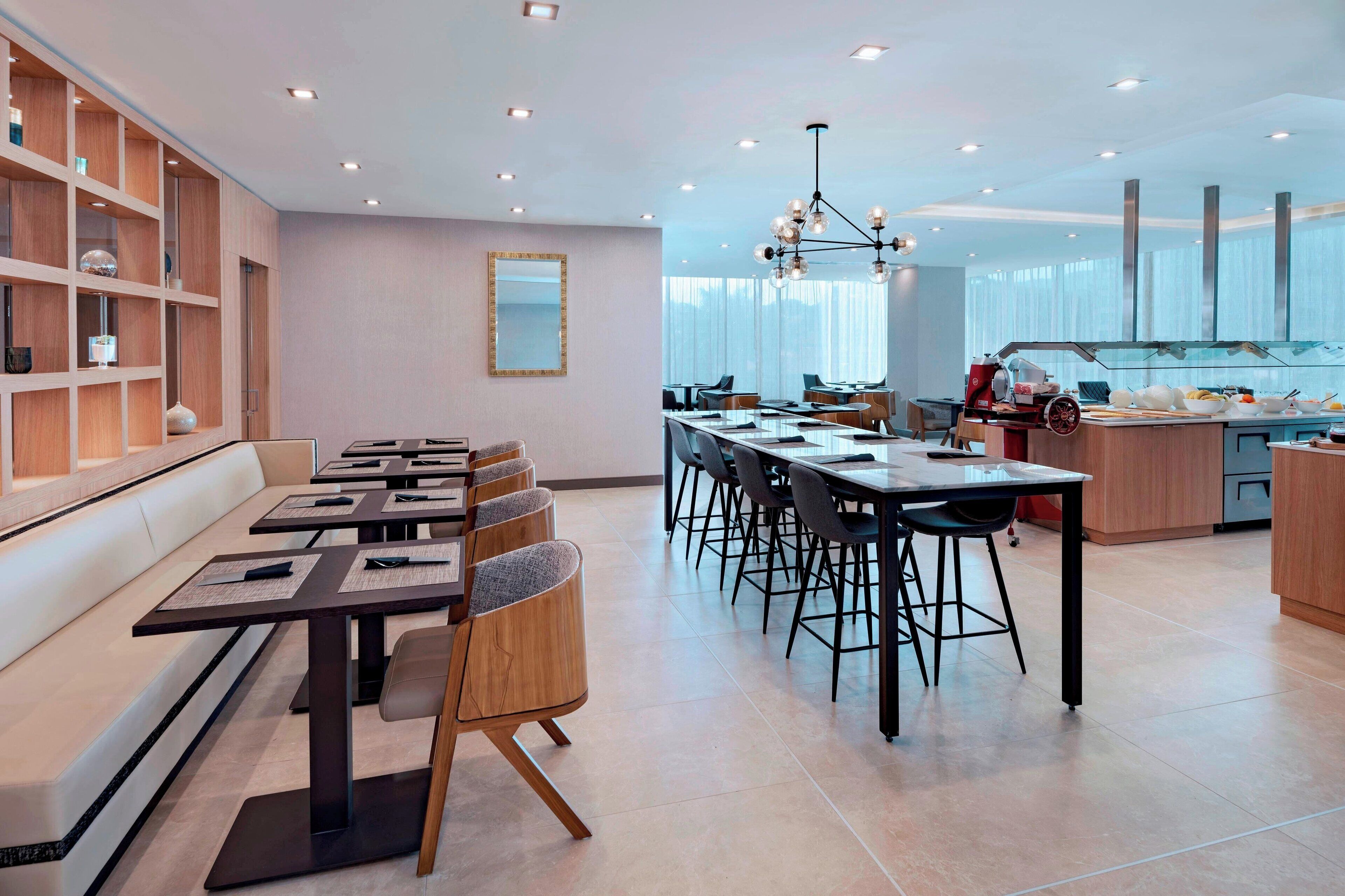 Een moderne eetruimte met lange tafels en stoelen, een keuken met een buffet en een groot raam dat natuurlijk licht binnenlaat.