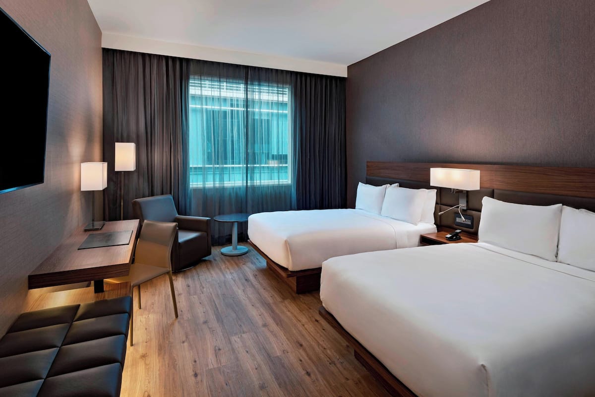 Quarto de hotel com duas camas, uma escrivaninha, uma cadeira, uma mesa pequena e uma televisão.