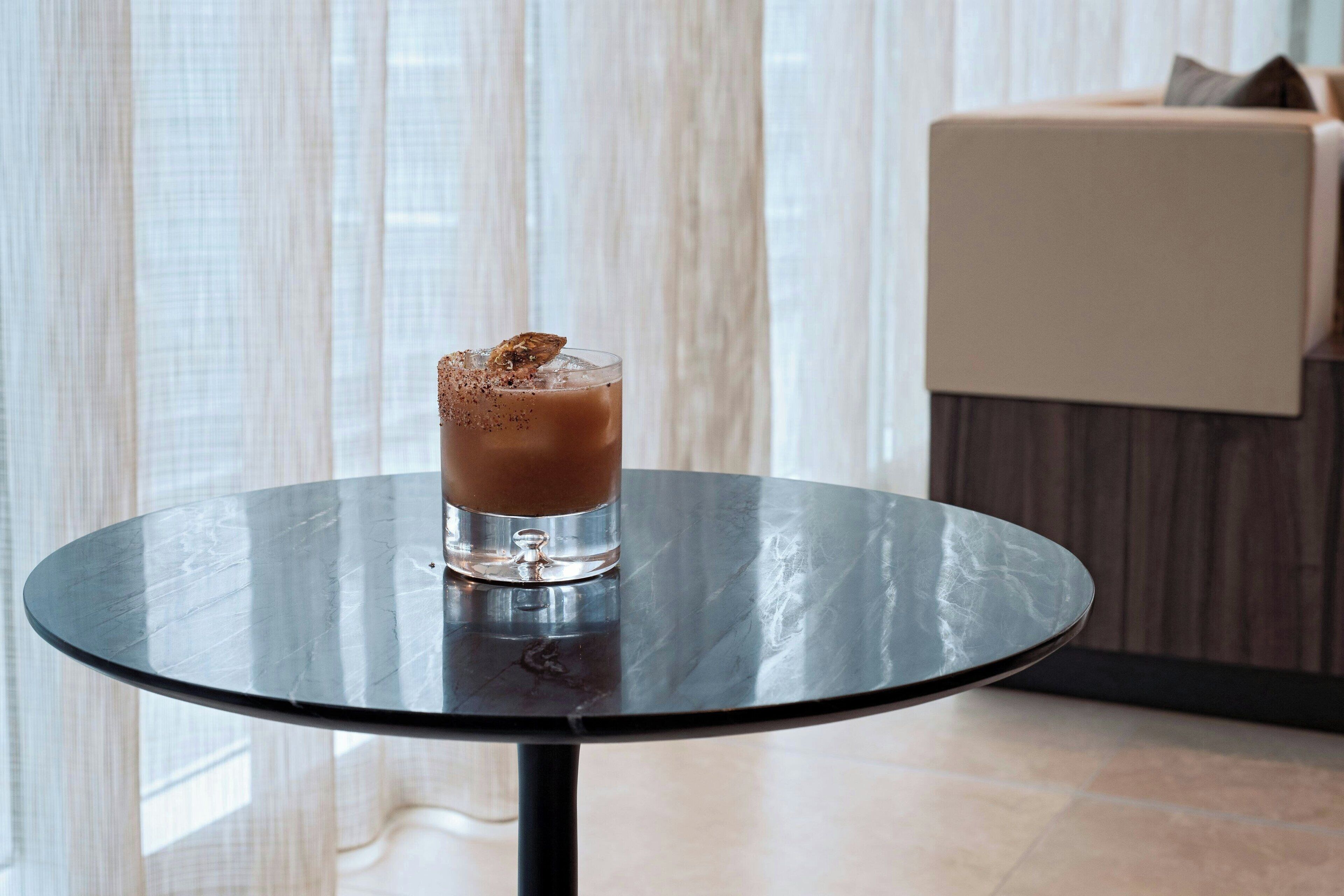 A glass of iced coffee on a round table with a metal frame, set against a backdrop of sheer curtains and a wooden wall.