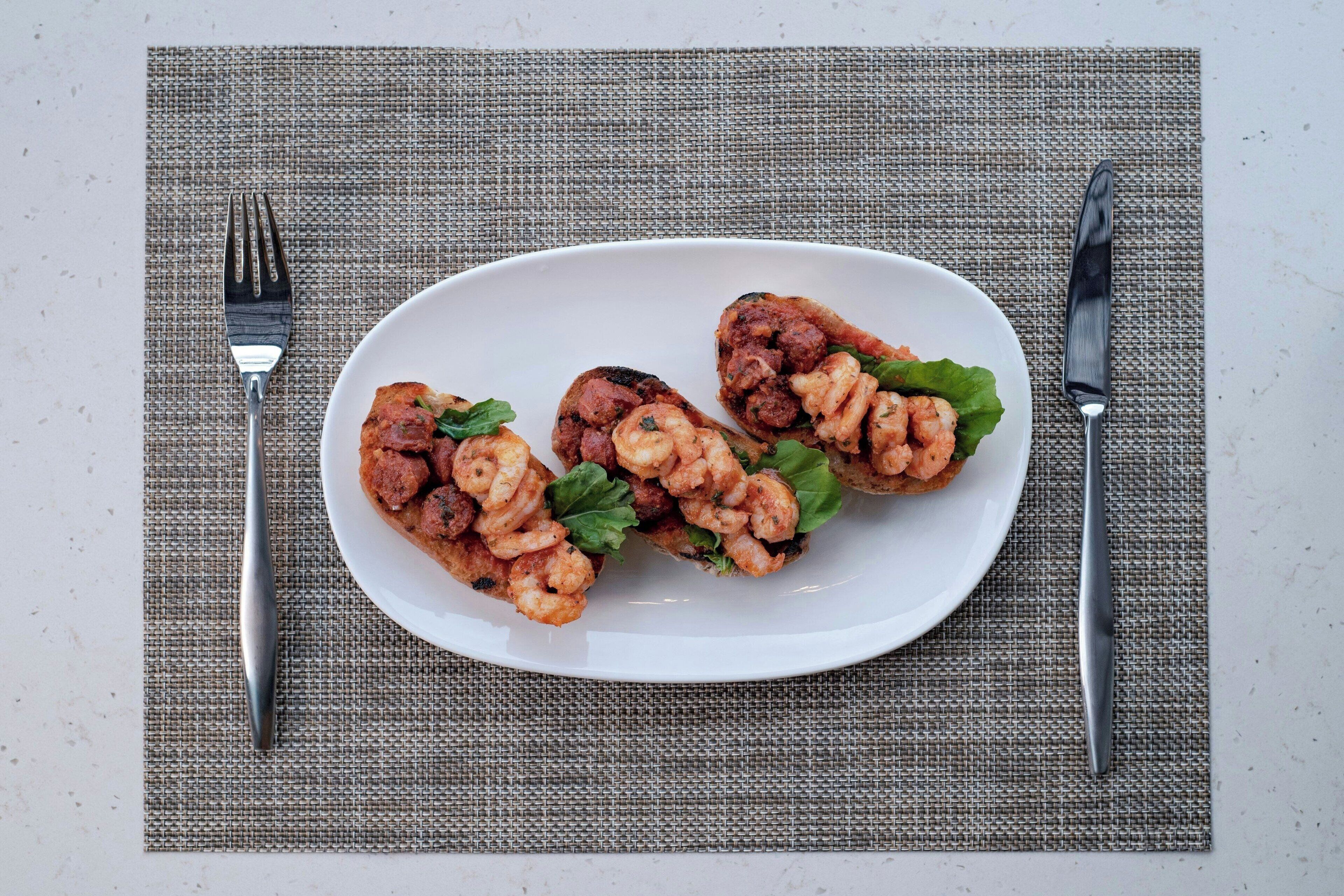 Un plato de camarones y salchicha sobre un plato blanco, colocado sobre un tapete texturizado con cubiertos a un lado.