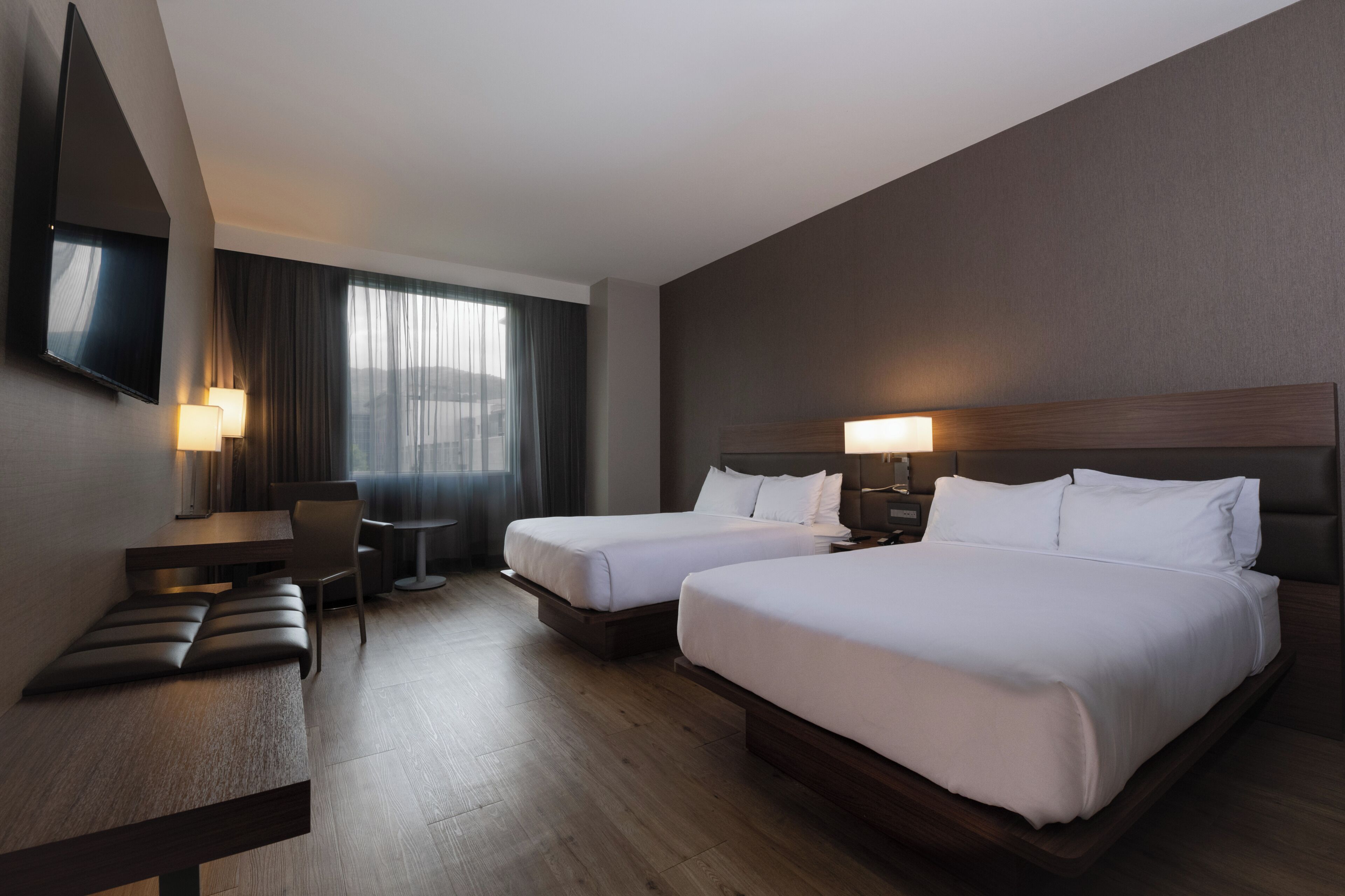 A hotel room with two beds, a desk, a chair, a TV, and a window with curtains.