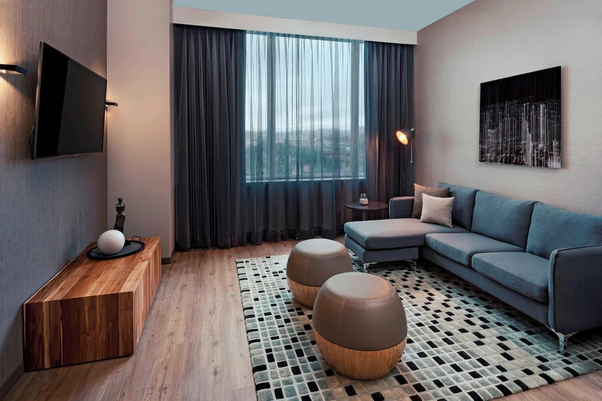 Uma sala de estar moderna com um sofá seccional, uma mesa de console de madeira, dois pufes, uma TV fixada na parede e uma grande janela com cortinas.