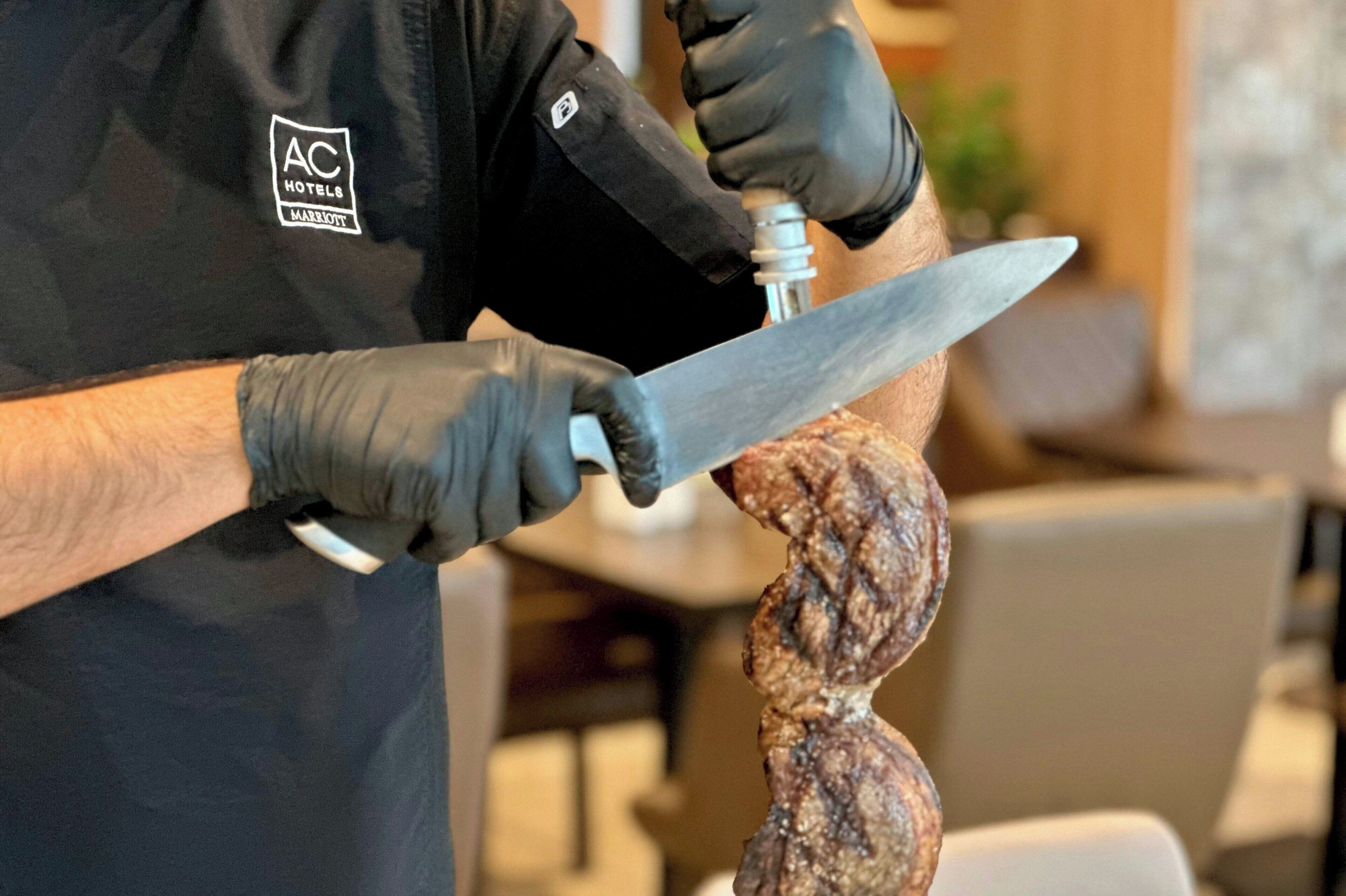 Una persona con delantal negro que lleva un logotipo, sosteniendo un cuchillo grande y un trozo de carne.