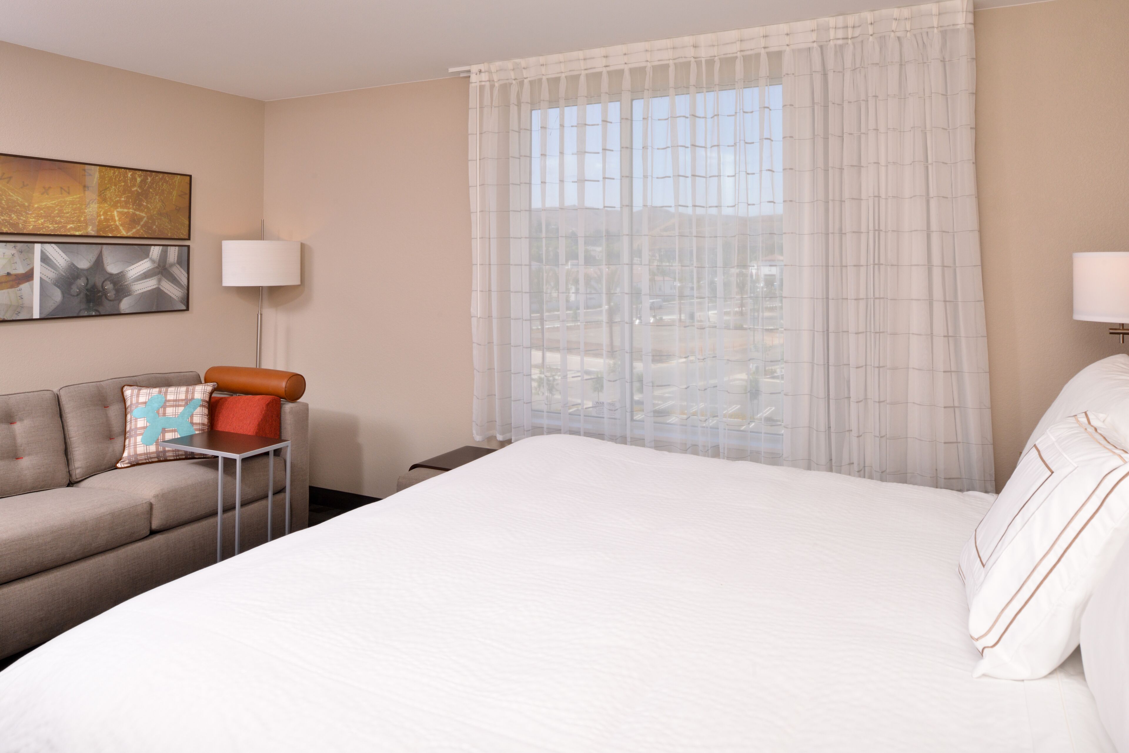 A hotel room with a large bed, a sofa, a small table, and a window with sheer curtains.