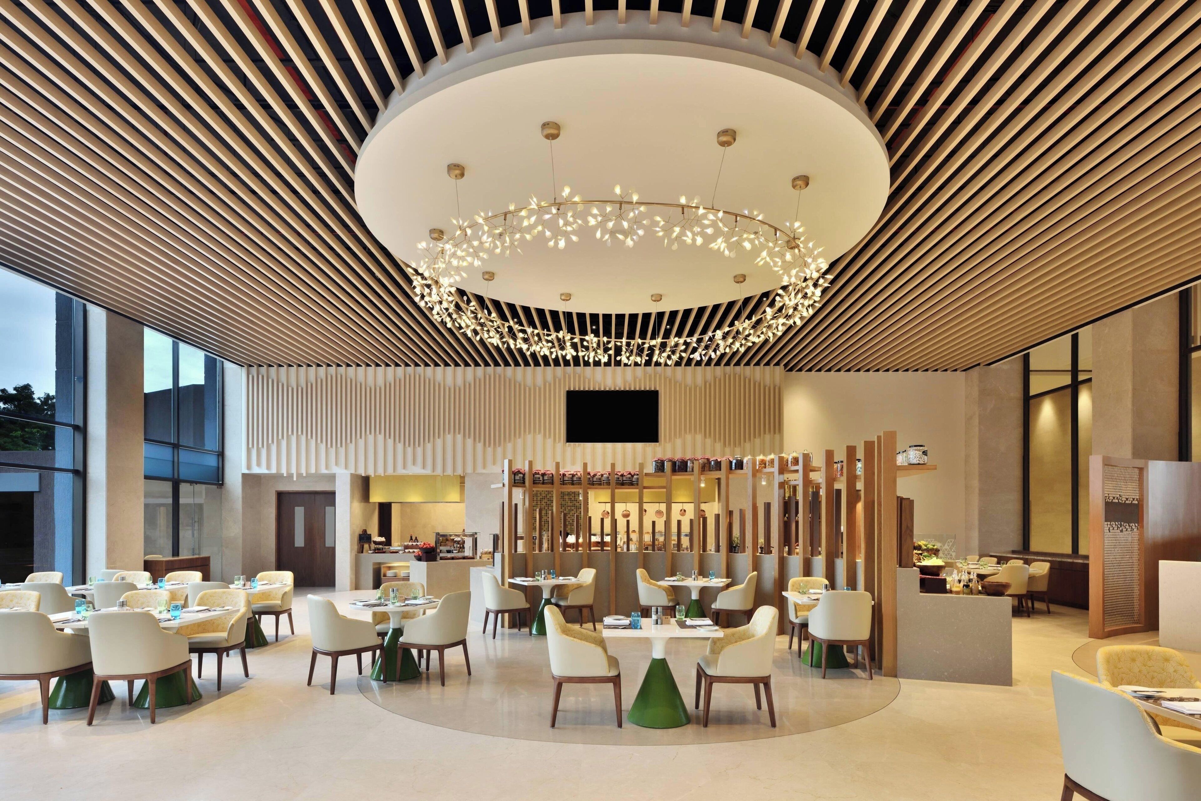A modern hotel lobby with round tables, comfortable seating, a central chandelier, and a large TV screen.