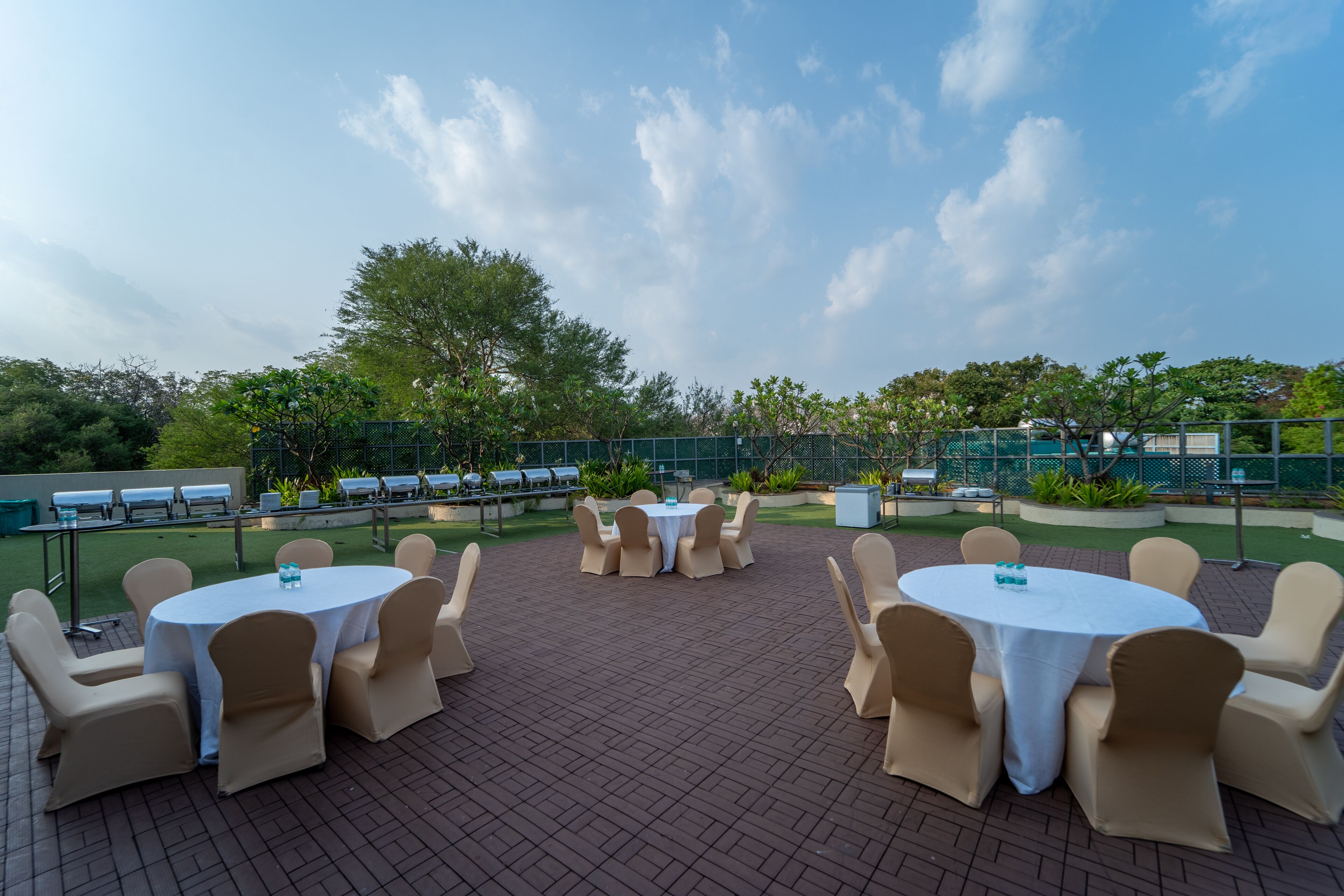 An outdoor patio area with round tables and chairs, surrounded by greenery and a clear sky.