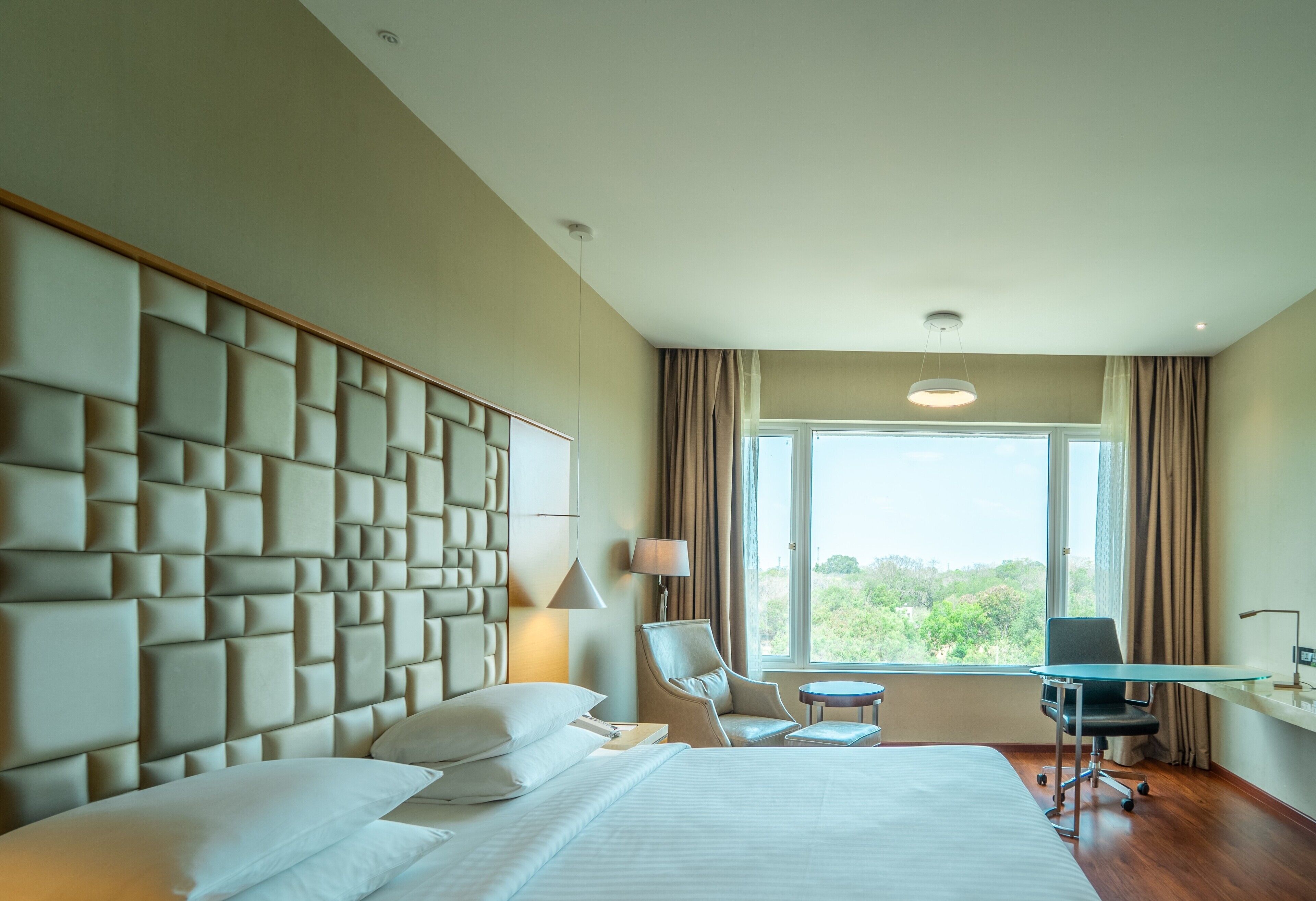 A hotel room with a large bed, a desk, a chair, and a view of greenery through the window.