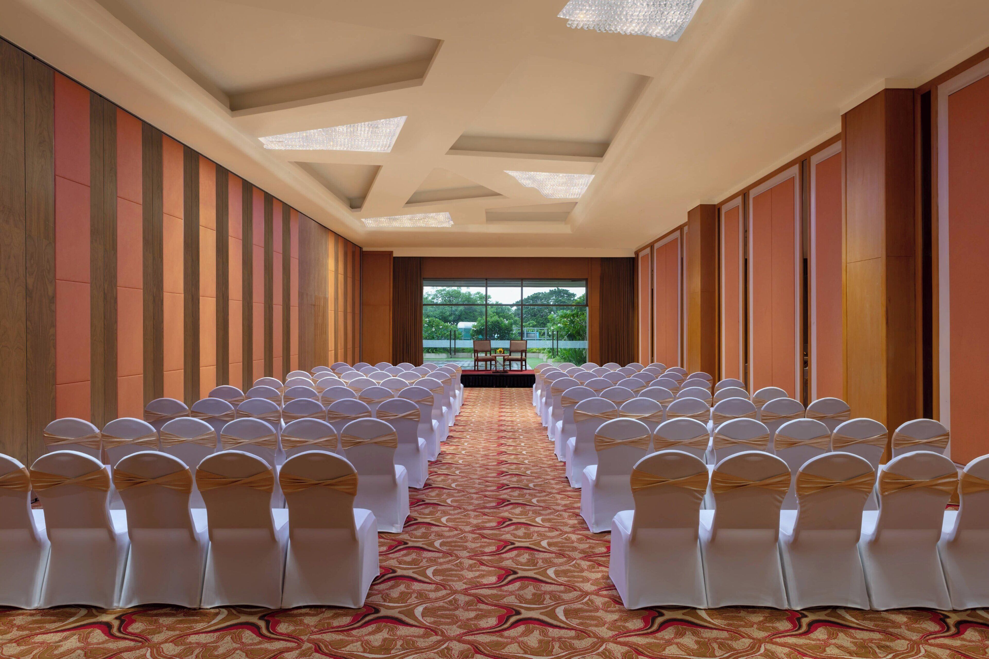 A conference room with rows of white chairs and a carpeted floor, leading to a stage with a view of greenery outside.