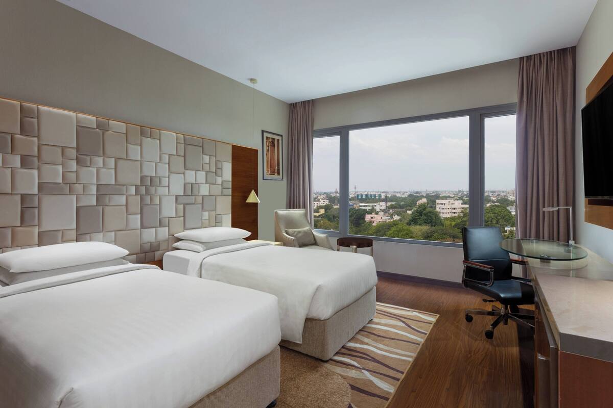 Ein Hotelzimmer mit zwei Betten, einem Schreibtisch, einem Stuhl, einem großen Fenster mit Stadtblick und einem modernen Wanddesign.