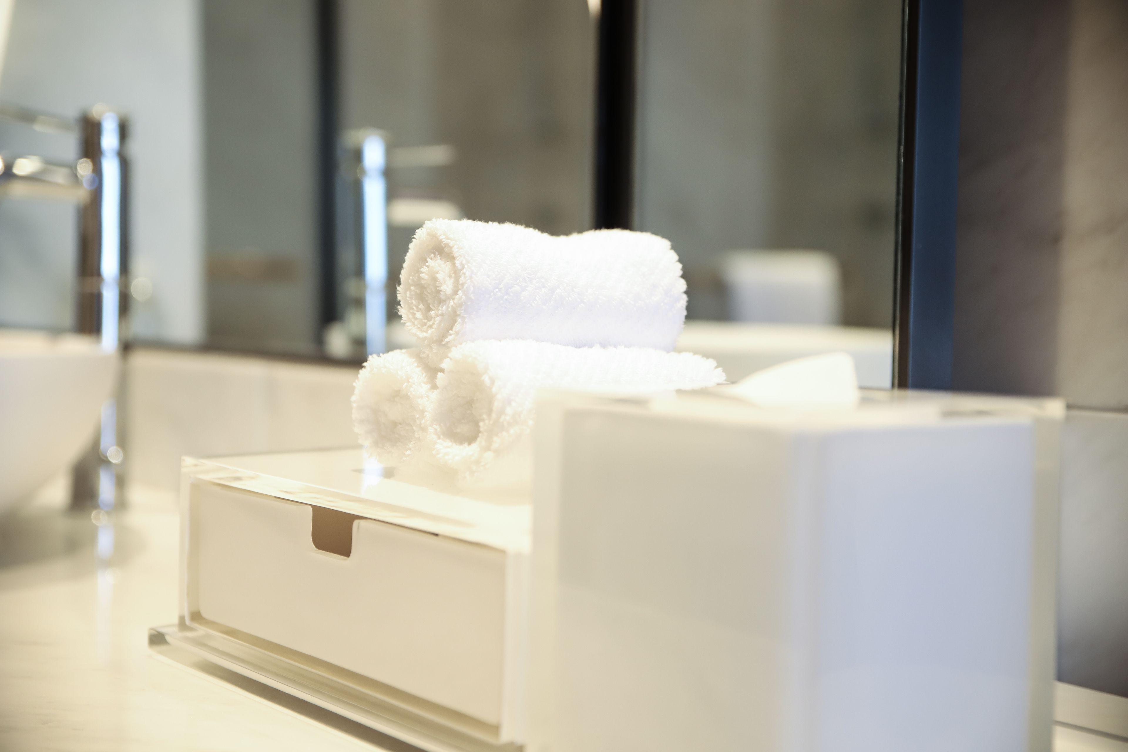 Een witte handdoek op een witte doos in een badkamer met een spiegel en een wastafel.