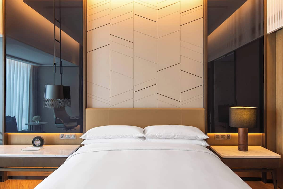 A modern hotel room with a large bed, a bedside table with a lamp, a mirror, and a view of the city through the window.