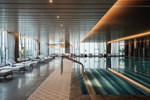 Un espace piscine intérieure moderne, doté de chaises longues et d’une paroi en verre.