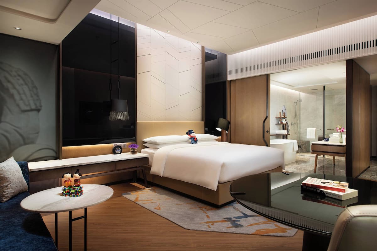 A modern hotel room with a large bed, a seating area with a small table, a large mirror, and a view of the bathroom.