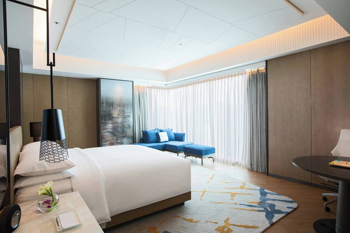 A modern hotel room with a large bed, a desk with a chair, a small table, and a sofa.