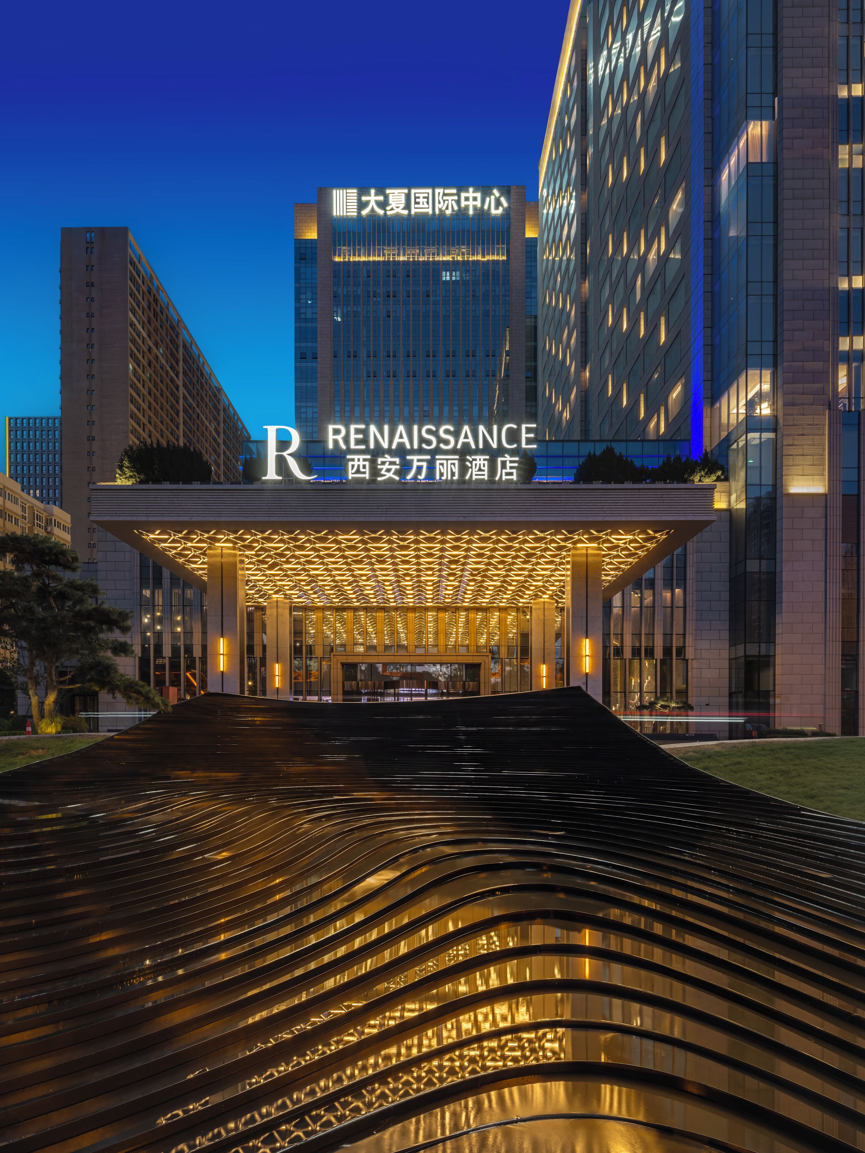 Una entrada de hotel moderna con un llamativo letrero 'Renaissance', fachada bien iluminada y un elemento acuático reflectante en primer plano.