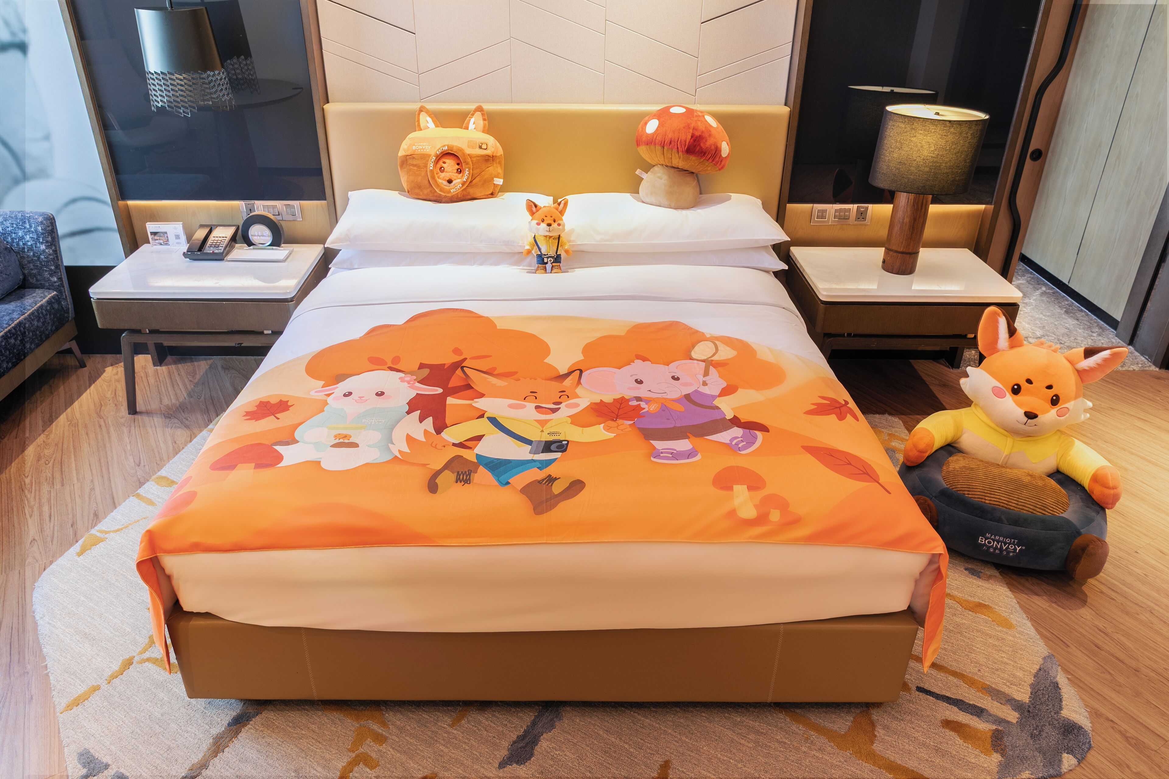 A hotel room with a large bed featuring a colorful autumn-themed bedspread, a small table with a lamp, a nightstand with a phone and a decorative item, and a plush toy on the bed.