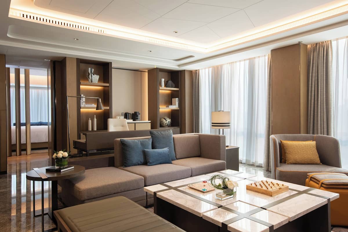 A modern hotel room with a large sofa, a coffee table, and a seating area with a chair. There is a bed in the background and a window with curtains.