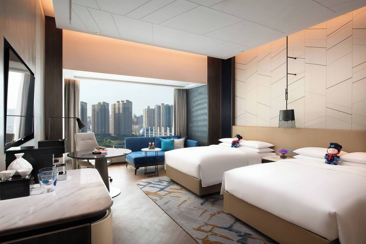 A hotel room with two beds, a sofa, a dining table, and a large window with a city view.
