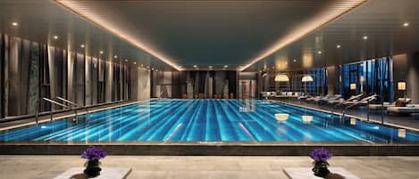 A modern indoor swimming pool with clear blue water, surrounded by lounge areas and a high ceiling with linear lighting.