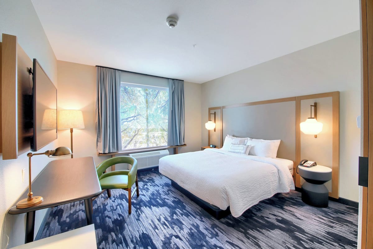 A hotel room with a bed, a desk, a chair, a window with curtains, and a lamp.