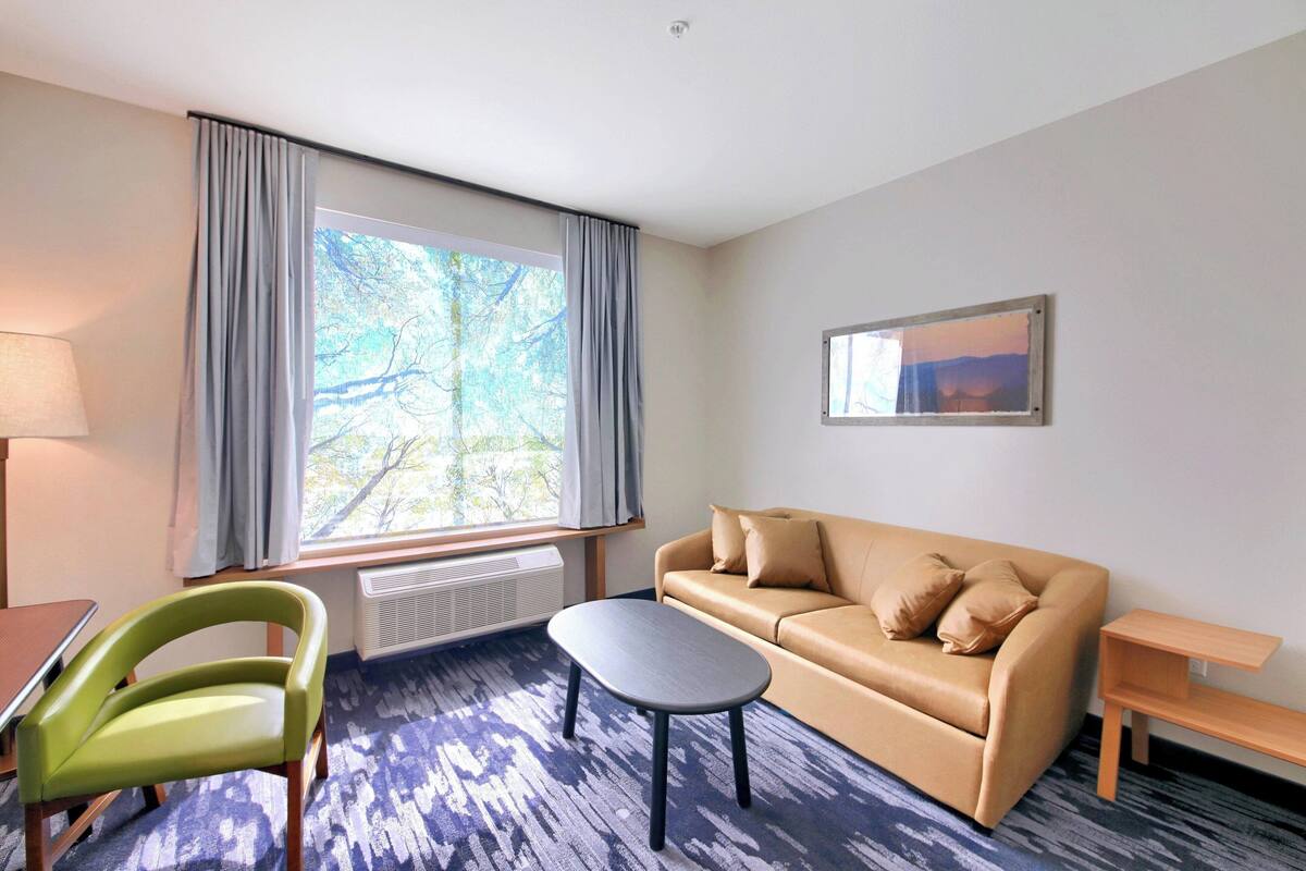 A modern hotel room with a sofa, a small table, a chair, and a large window with curtains.