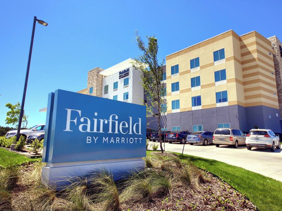 A Fairfield by Marriott hotel sign with a parking lot and a multi-story building in the background.