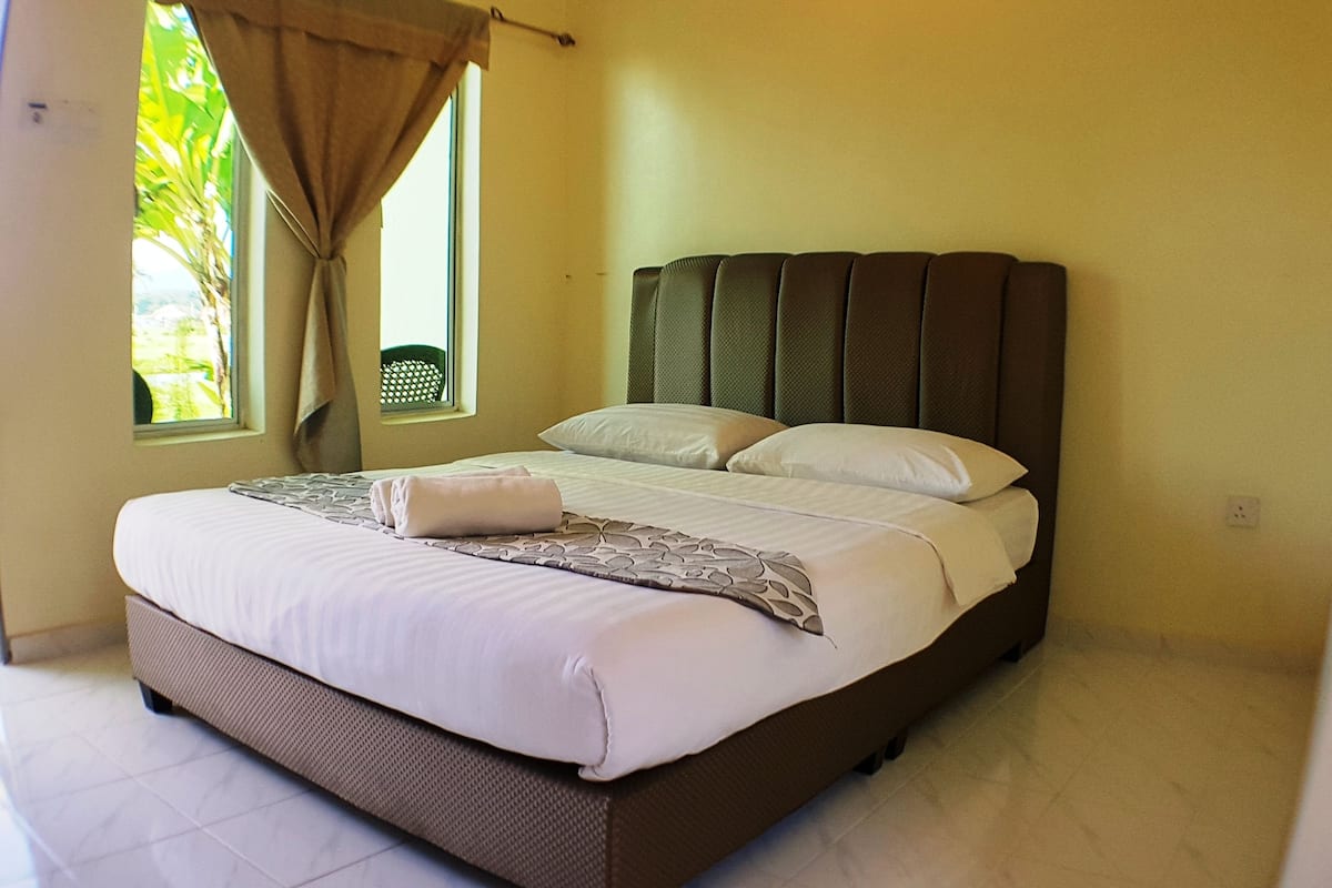 A neatly made bed with white linens and a patterned throw, a window with a view of greenery, and a brown headboard.