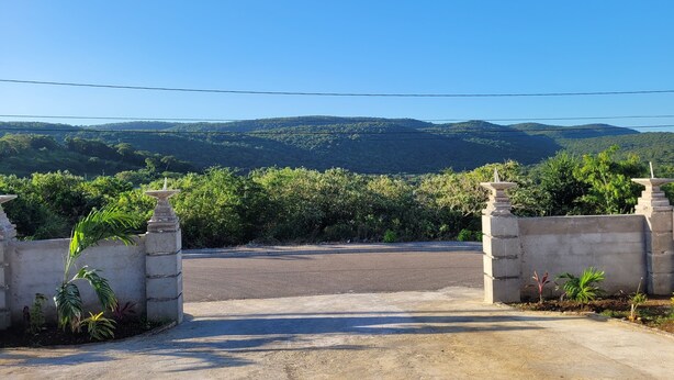 A paved entrance with stone pillars, leading to a road with a view of green hills and a clear blue sky.
