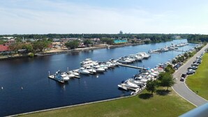 A marina with multiple boats docked along a river, surrounded by greenery and buildings.
