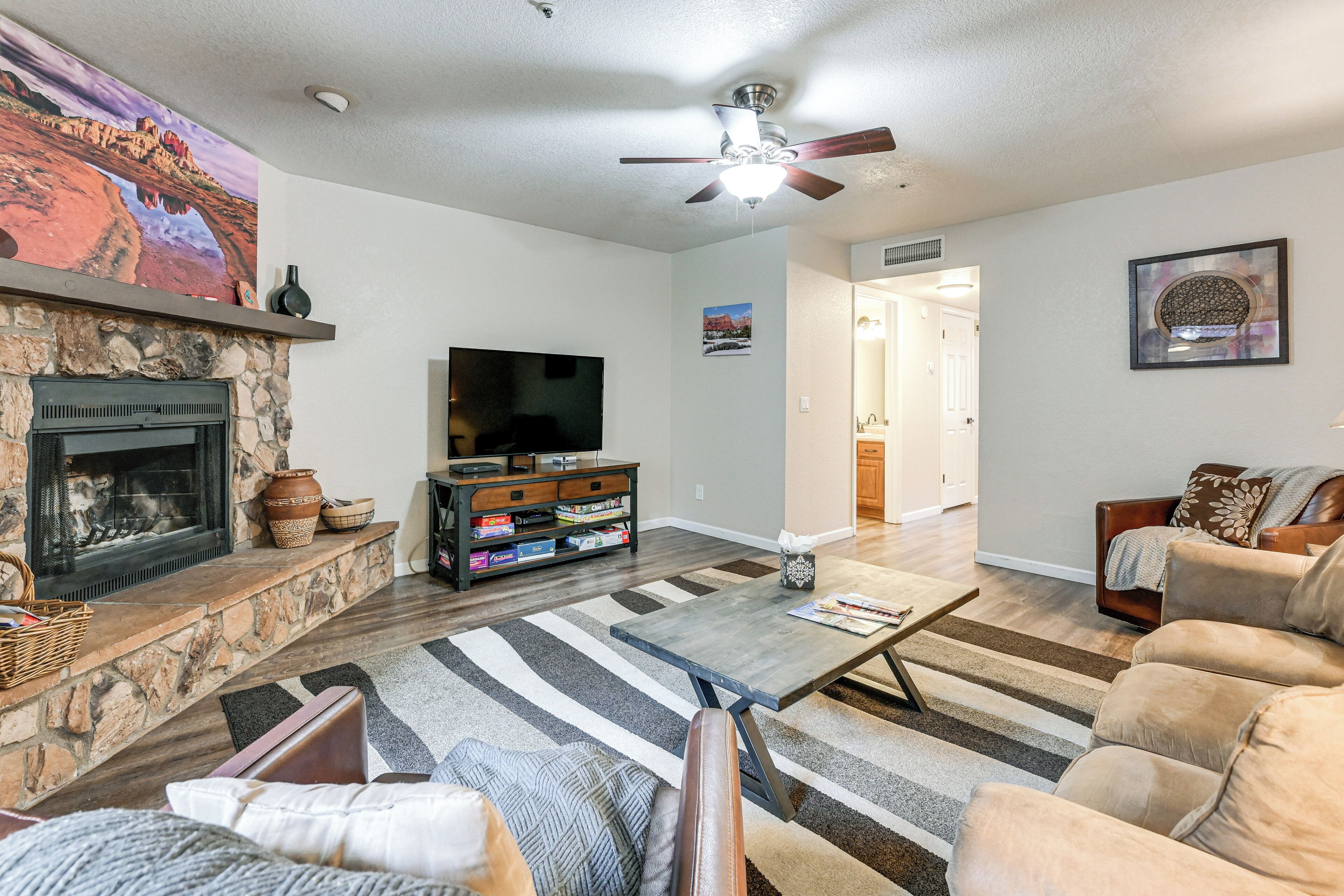 A living room with a stone fireplace, a flat-screen TV, a ceiling fan, and a large rug.