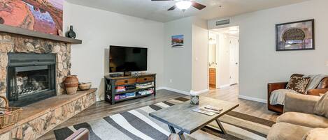 A living room with a stone fireplace, a flat-screen TV, a ceiling fan, and a large rug.