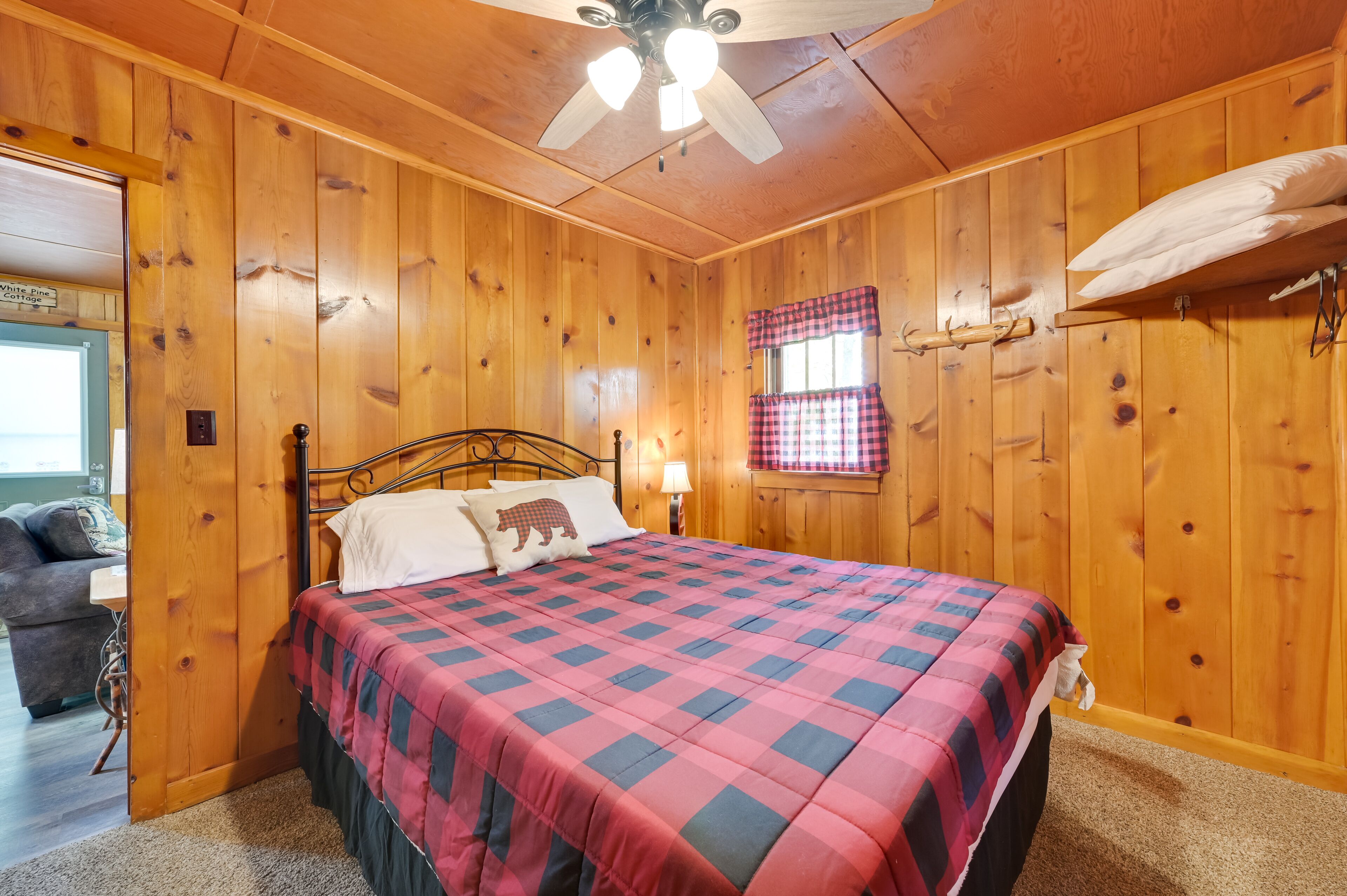 A wooden-paneled bedroom with a bed, a nightstand, a lamp, and a window with plaid curtains.