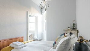 Ein ordentlich bezogenes Bett mit weißen Leinen und einer gelben Tagesdecke, eine Nachttischlampe und ein kleines Regal mit einer Uhr und einer Topfpflanze.